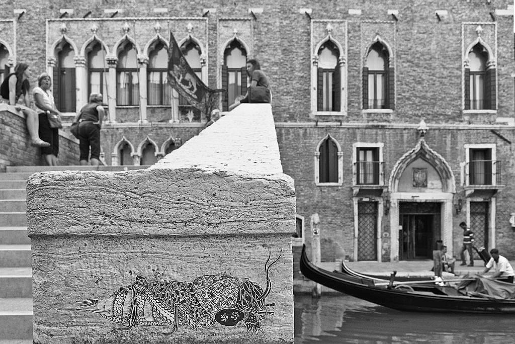 Stencil di un grillo su di un ponte a Venezia, con gente sul punte e sullo sfondo un palazzo gotico con una  gondola d'avanti.